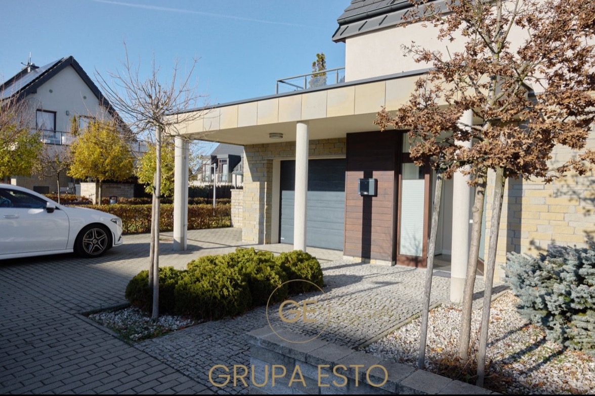 Nowoczesny dom jednorodzinny z podjazdem wyłożonym kostką, garażem w bryle budynku i starannie zaprojektowanym ogrodem z krzewami i młodymi drzewami.