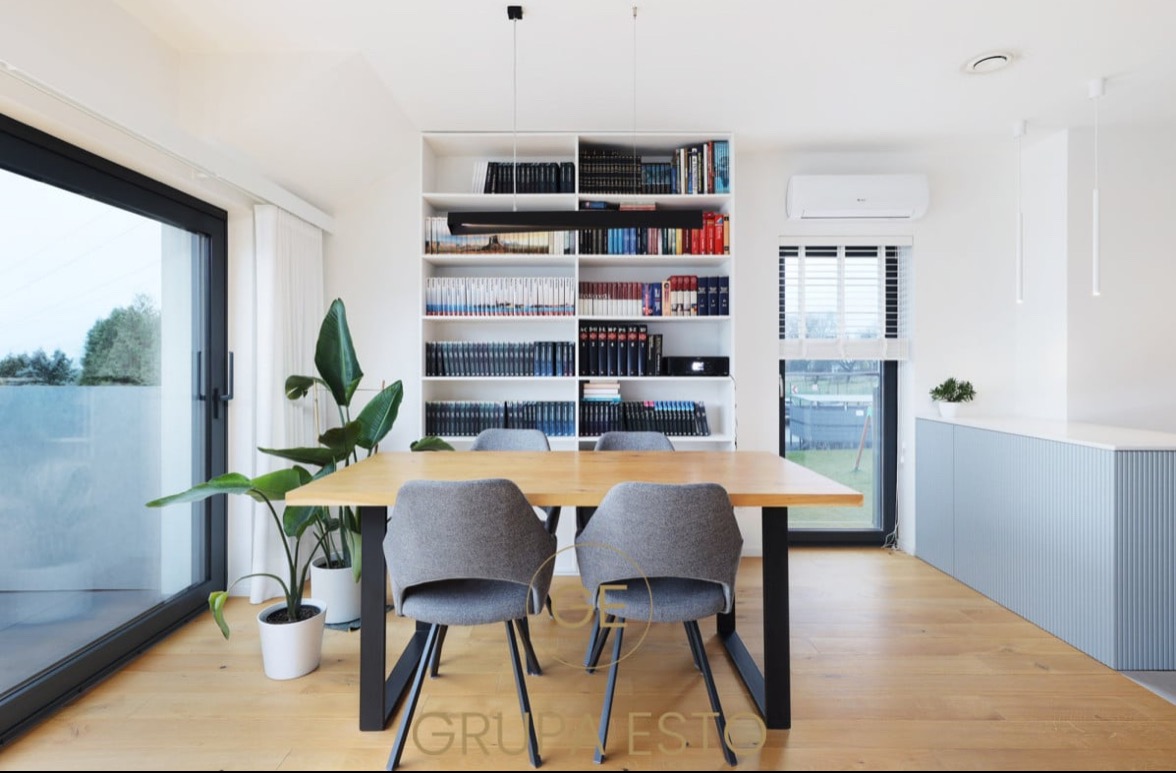 Jasna jadalnia z drewnianym stołem, szarymi krzesłami i regałem z książkami, widok na ogród przez duże okno, nowoczesne oświetlenie punktowe.