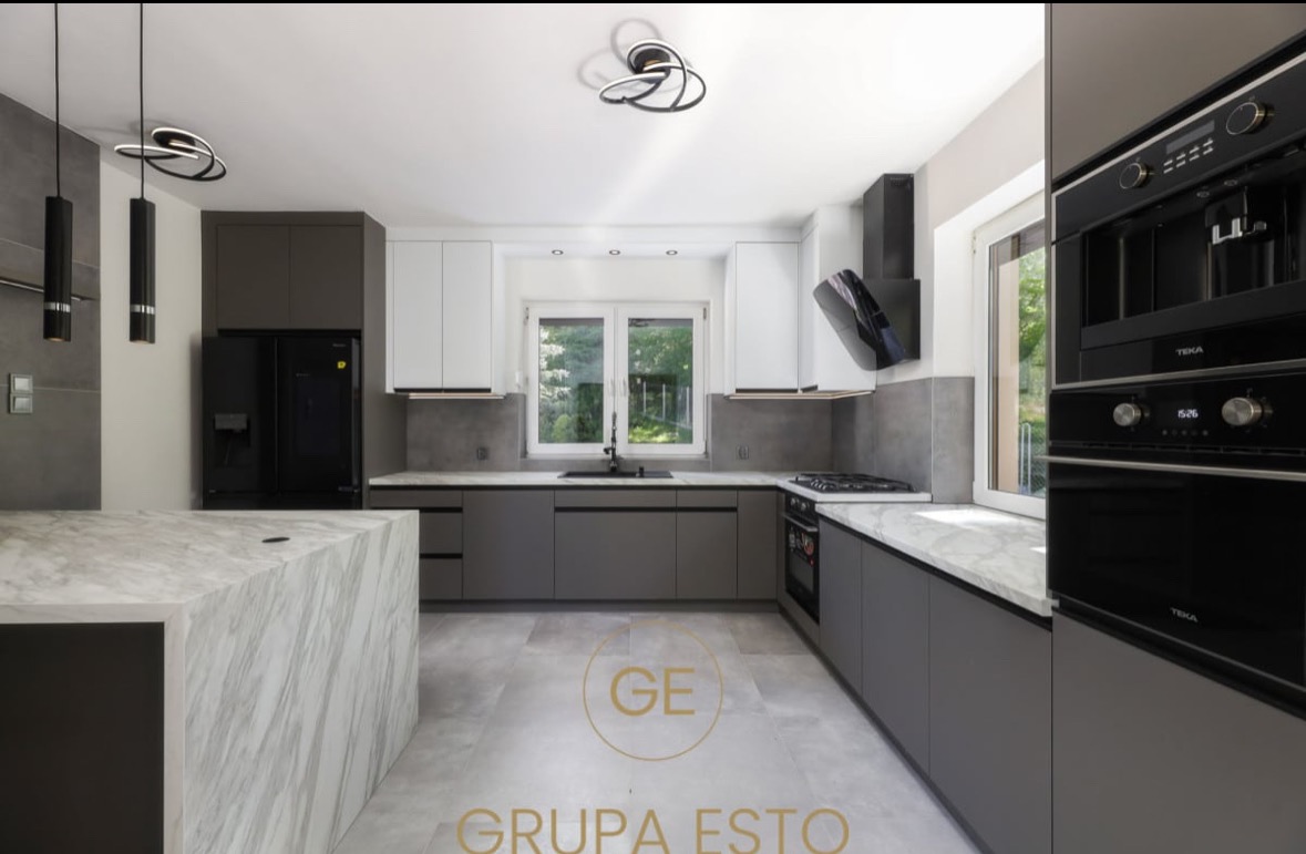 Nowoczesna kuchnia z szarymi szafkami, białymi górnymi szafkami i marmurowym blatem wyspy, oświetlona designerskimi lampami wiszącymi i sufitowymi.