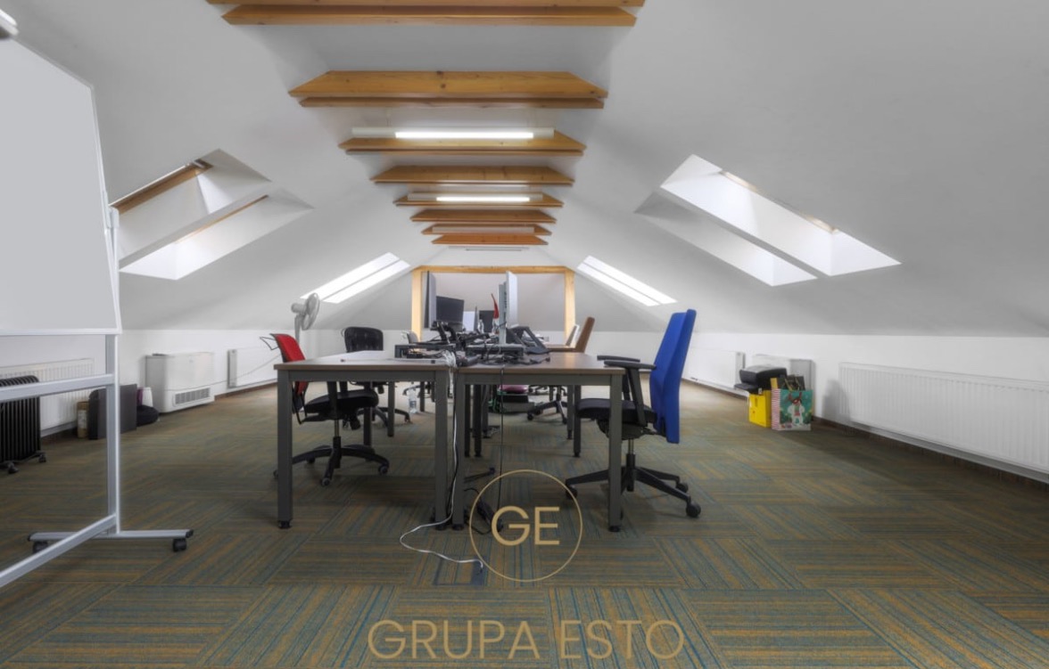 Przestronne biuro na poddaszu z oknami dachowymi, wyposażone w biurka, krzesła (jedno niebieskie), tablicę suchościeralną i grzejniki, z widocznym logo 'Grupa Esto' na pierwszym planie.