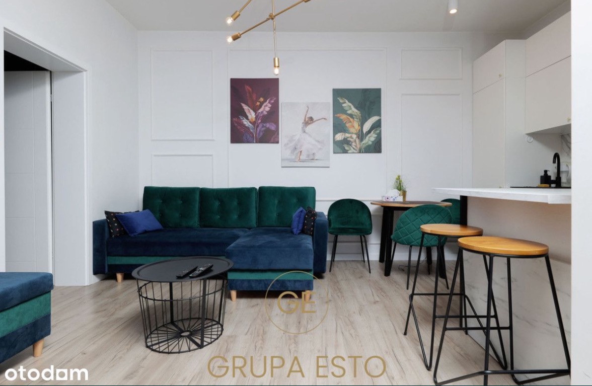 Wnętrze salonu z aneksem kuchennym: ciemnozielona sofa narożna, okrągły stolik kawowy z pilotami, drewniane stołki barowe, białe ściany z dekoracyjnymi panelami i trzema obrazami.