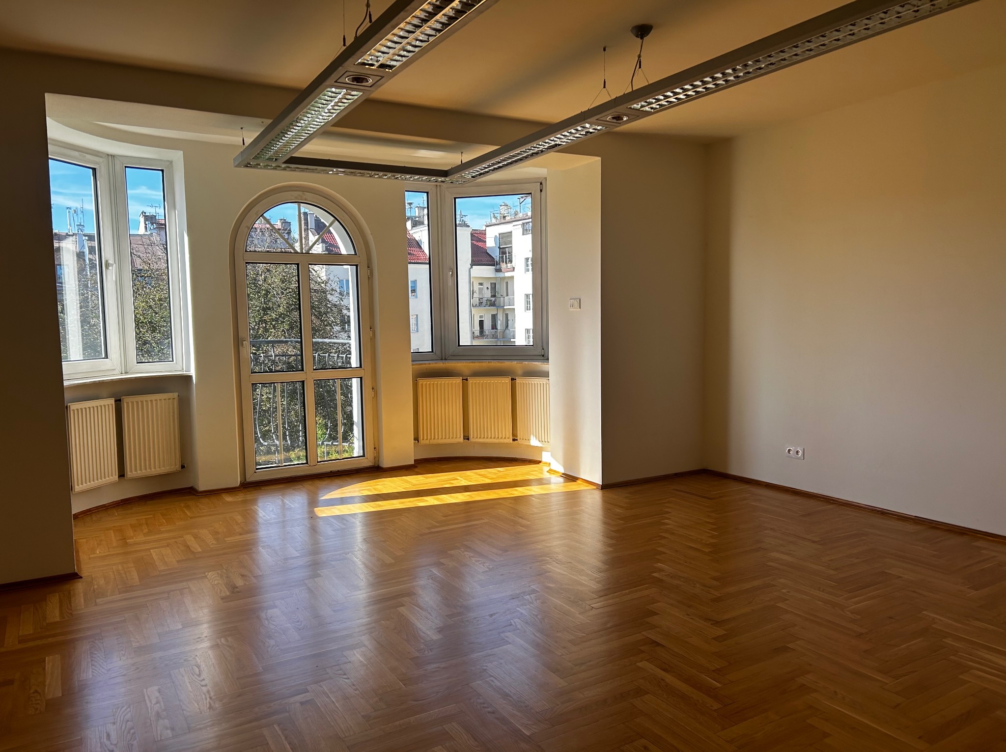 Puste, jasne pomieszczenie z parkietem w jodełkę, trzema oknami z widokiem na budynki i drzewa, oraz nowoczesnym oświetleniem sufitowym.