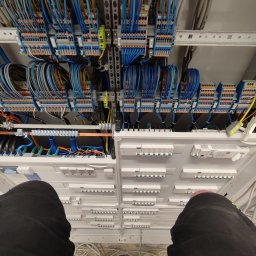 Alek-Tech Aleksandra Żmuda - Szafa sterownicza z licznymi przewodami, złączkami i zabezpieczeniami, widok z góry z fragmentem sylwetki osoby w czarnych spodniach w dolnej części kadru.