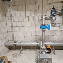 Alek-Tech Aleksandra Żmuda - Szara rura z elementami armatury, w tym niebieską skrzynką i pomarańczowym silnikiem, zamontowana w surowym pomieszczeniu z bloków betonowych, z widocznymi metalowymi korytkami kablowymi...