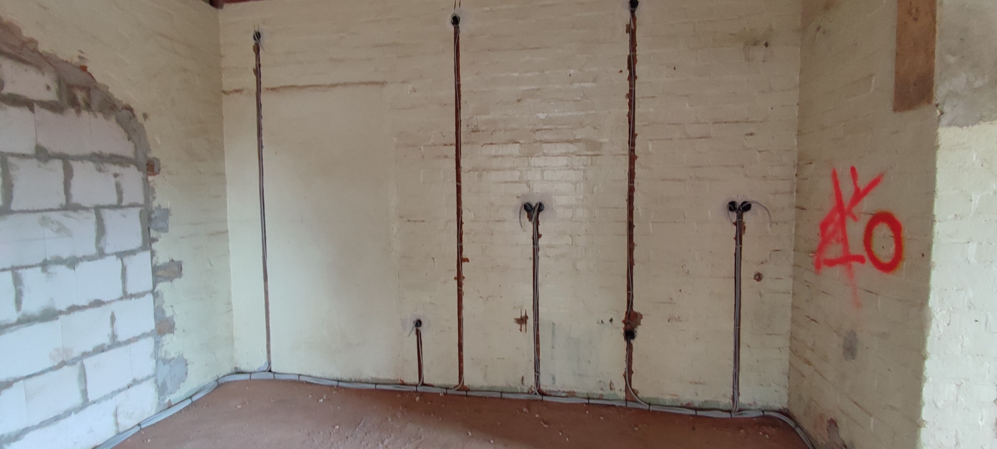 Ściana z instalacją elektryczną w trakcie montażu; widoczne kable w bruzdach, puszki pod gniazdka i przewody przy podłodze. Na ścianie czerwone graffiti.