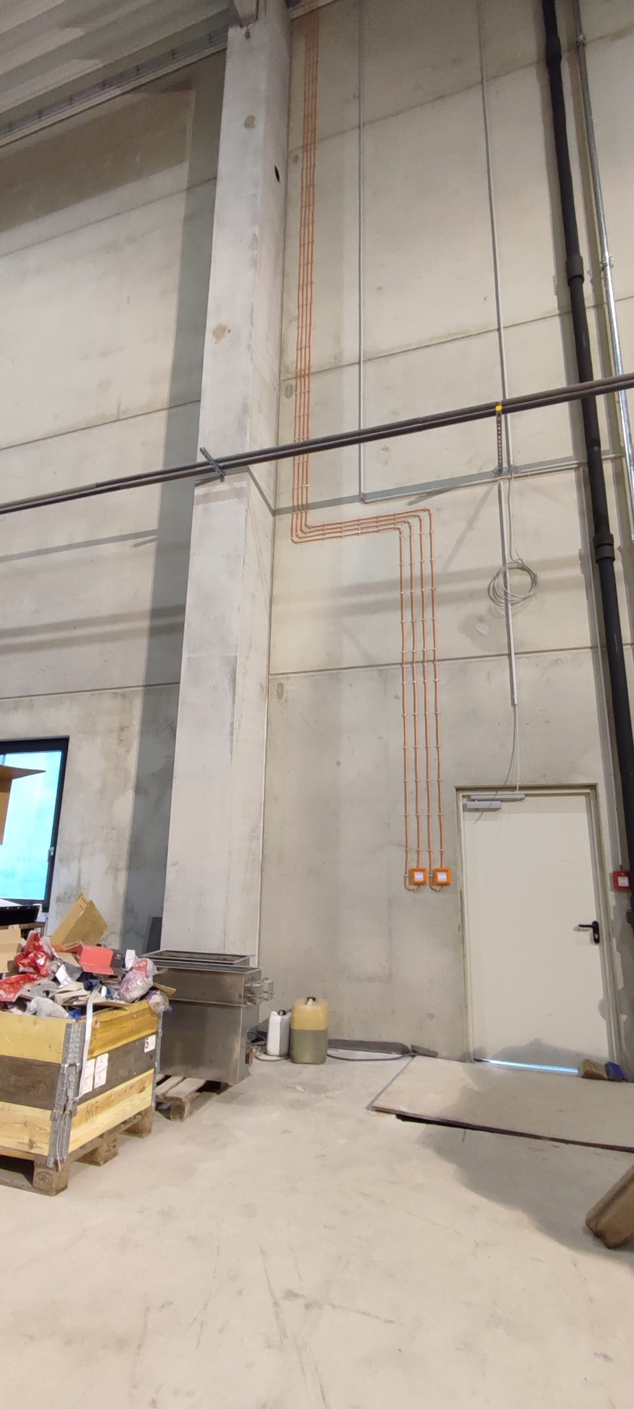 Surowe wnętrze hali z widocznymi, nowo poprowadzonymi przewodami elektrycznymi w pomarańczowych rurkach, zamontowanymi na betonowej ścianie. Przyłącza elektryczne z pomarańczowymi puszkami...