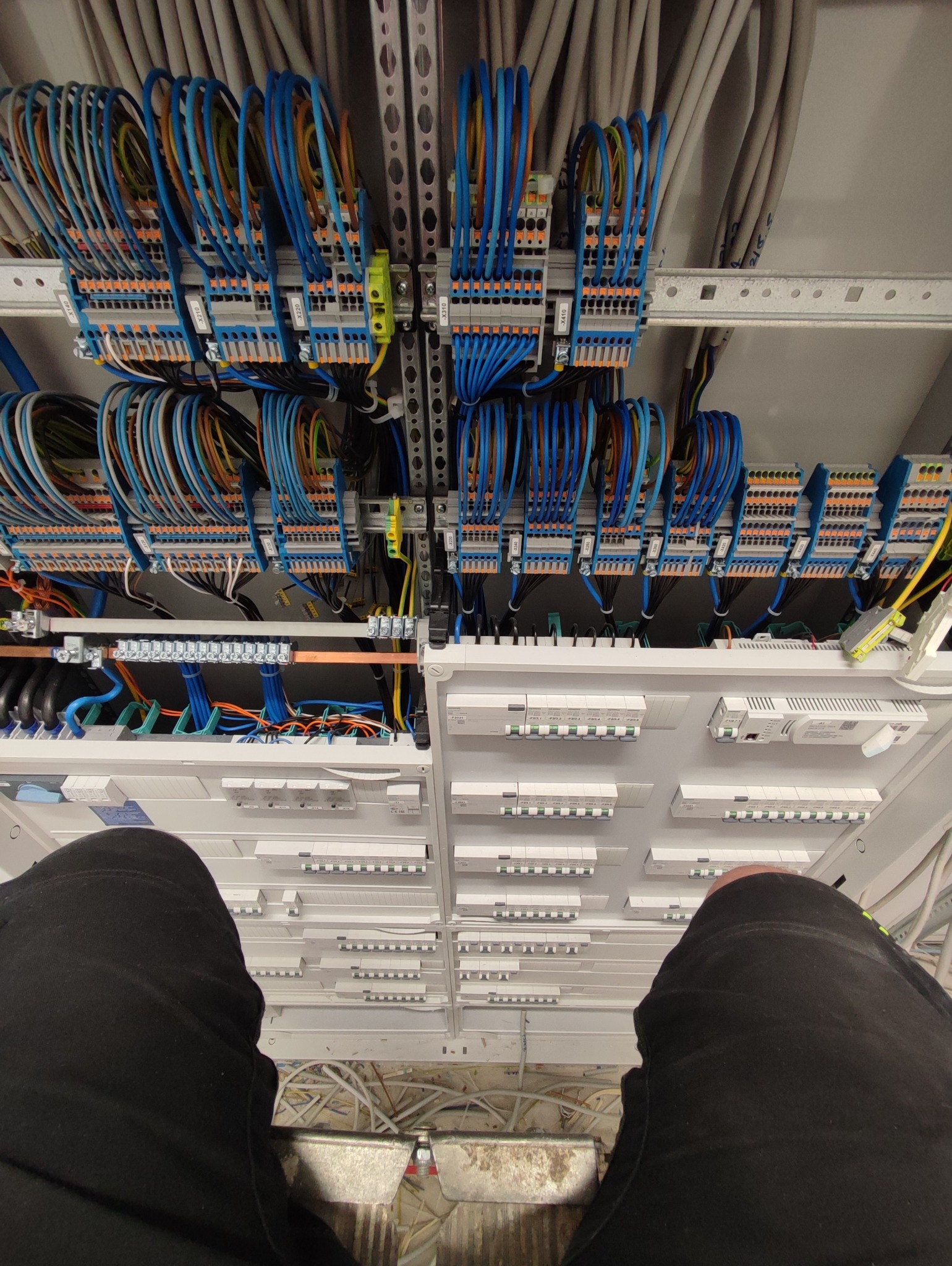 Szafa sterownicza z licznymi przewodami, złączkami i zabezpieczeniami, widok z góry z fragmentem sylwetki osoby w czarnych spodniach w dolnej części kadru.