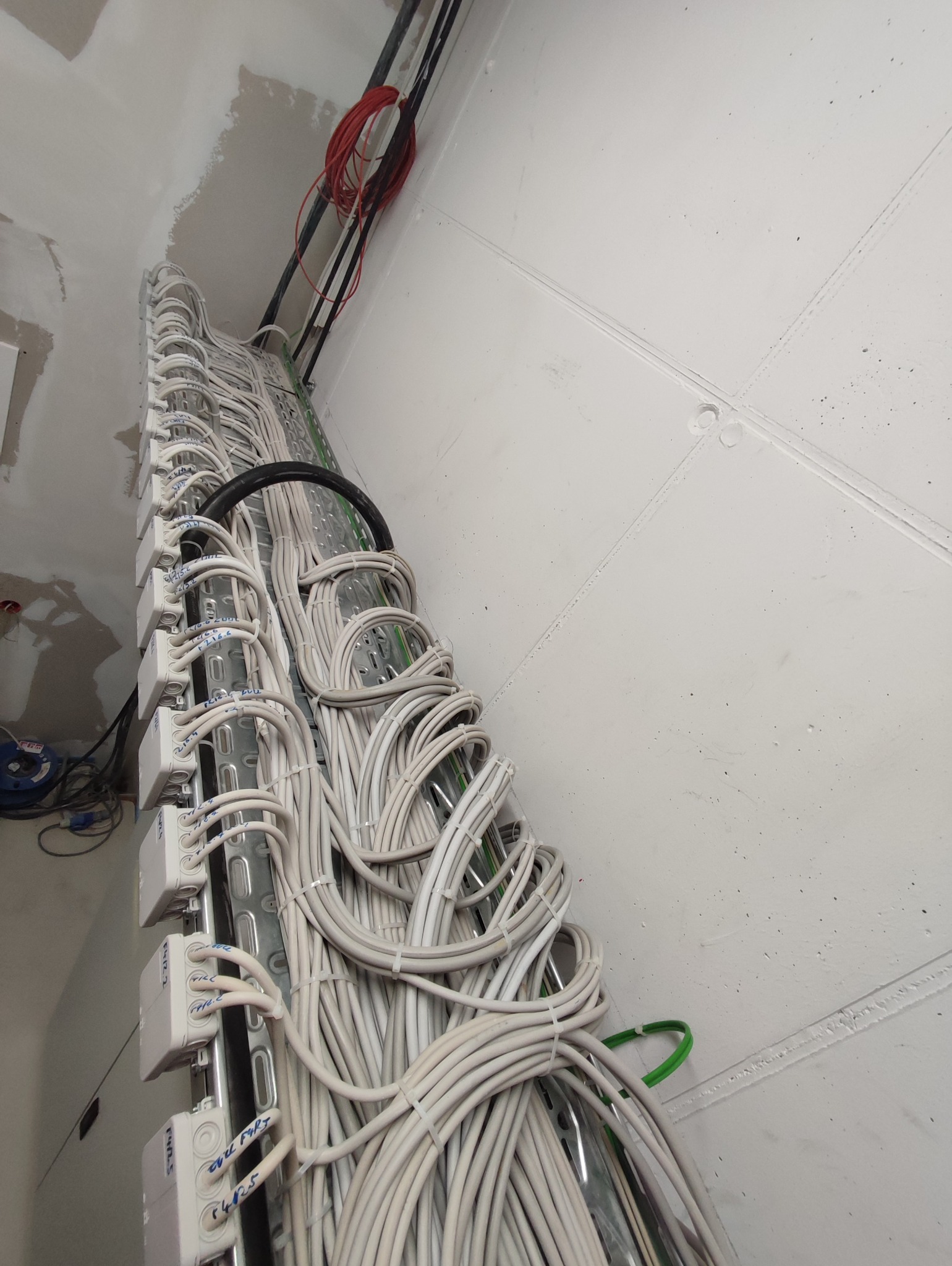 Rzędy białych kabli elektrycznych, ułożone w metalowej korycie pod sufitem w surowym stanie, z widocznymi puszkami połączeniowymi z odręcznymi oznaczeniami.