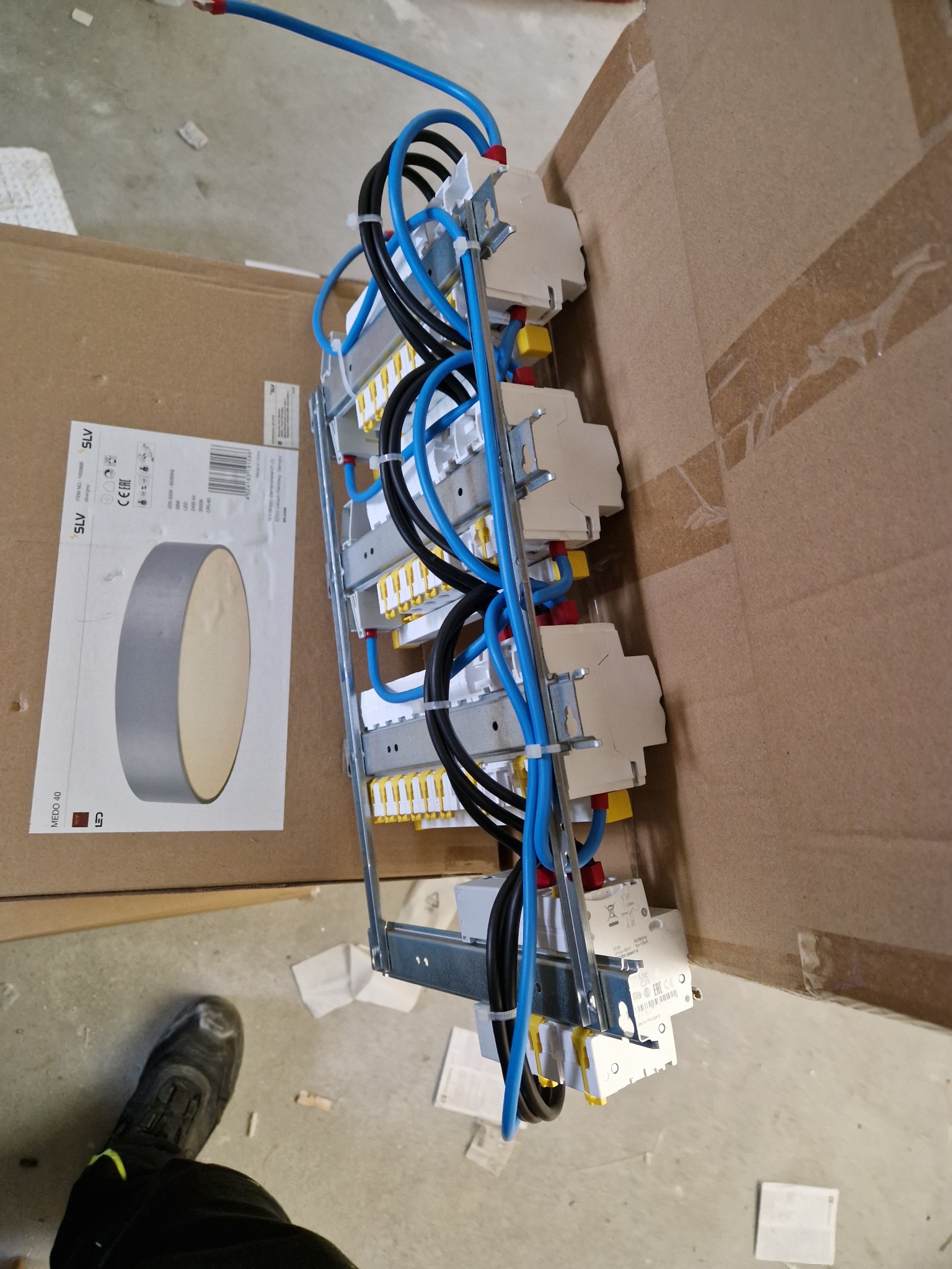 Rozdzielnica elektryczna z zamontowanymi bezpiecznikami i przewodami w kolorach niebieskim i czarnym, widoczna na tle kartonowych opakowań, z fragmentem buta osoby wykonującej prace w dolnym lewym...