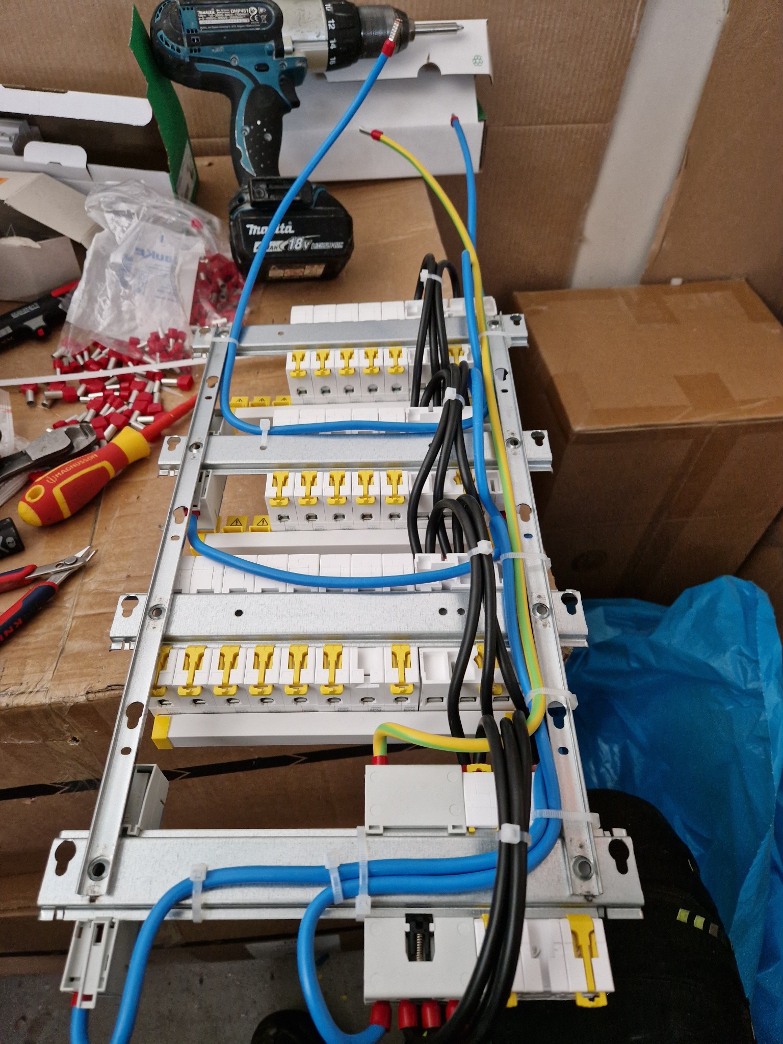 Rozdzielnica elektryczna w trakcie montażu, widoczne przewody w kolorach niebieskim, czarnym i żółto-zielonym, zabezpieczone opaskami zaciskowymi, obok wkrętarka akumulatorowa Makita i narzędzia.