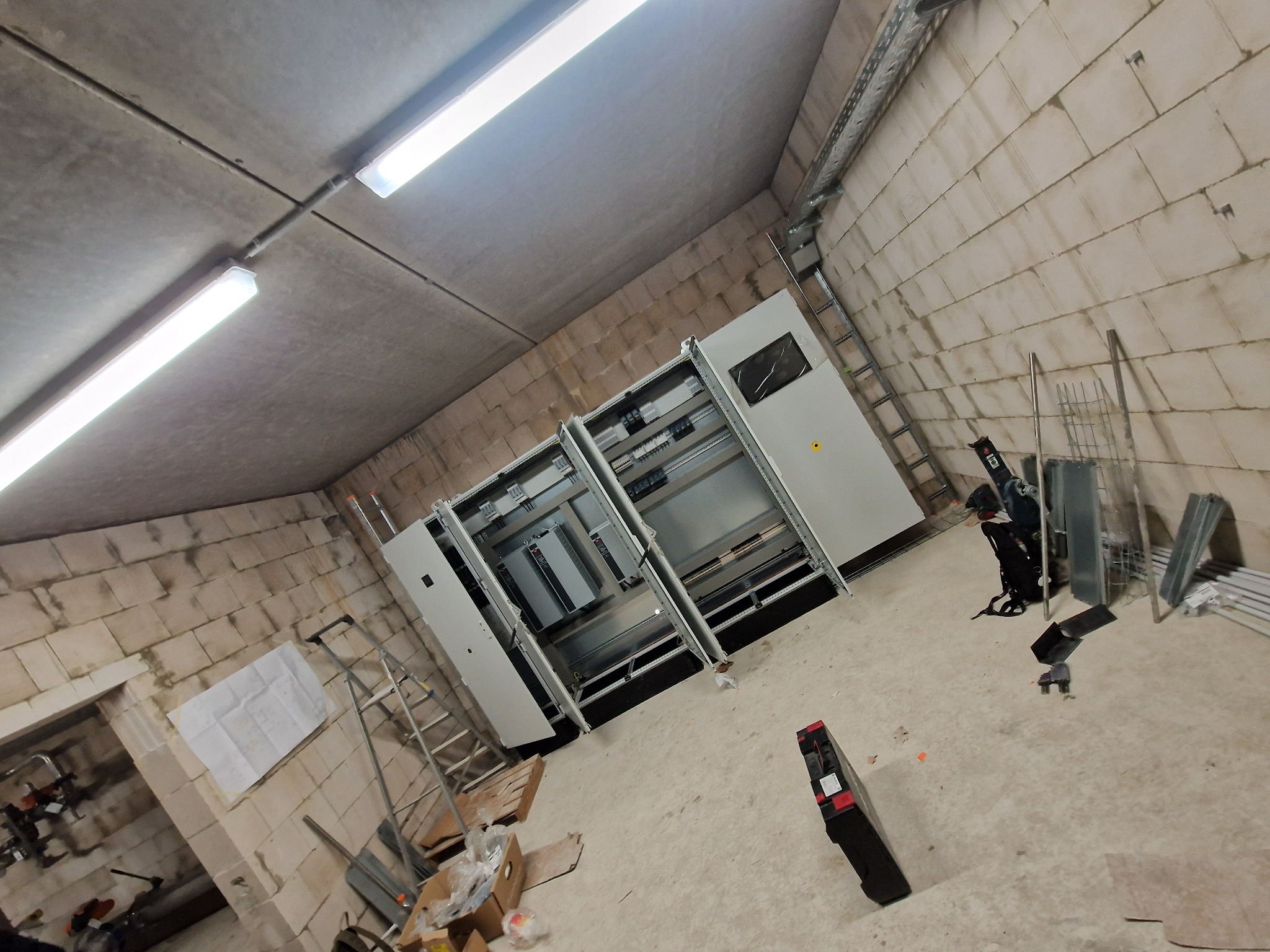 Rozdzielnica elektryczna w trakcie montażu w surowym pomieszczeniu z widocznym oświetleniem LED, drabina i narzędzia.