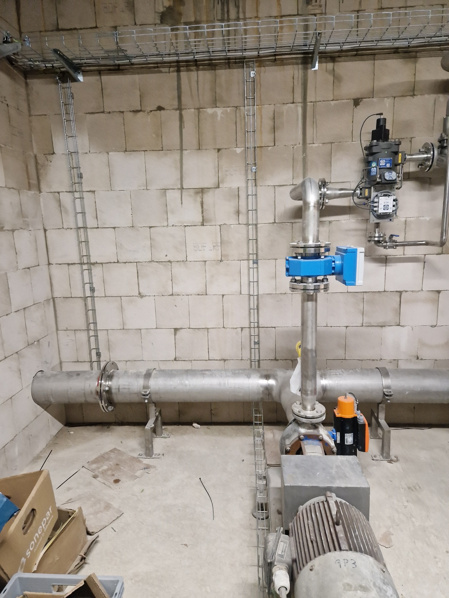 Szara rura z elementami armatury, w tym niebieską skrzynką i pomarańczowym silnikiem, zamontowana w surowym pomieszczeniu z bloków betonowych, z widocznymi metalowymi korytkami kablowymi...