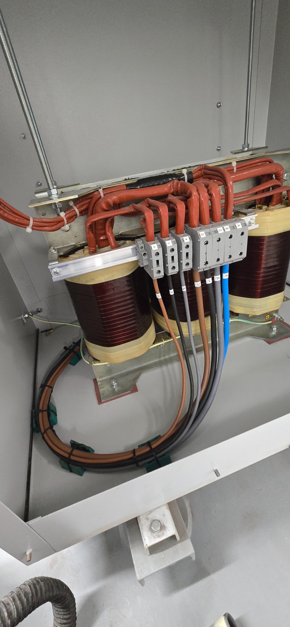 Transformator w szafie rozdzielczej z podłączonymi przewodami o różnych kolorach, widoczne złącza i mocowania. Element instalacji energetycznej w szarej obudowie.