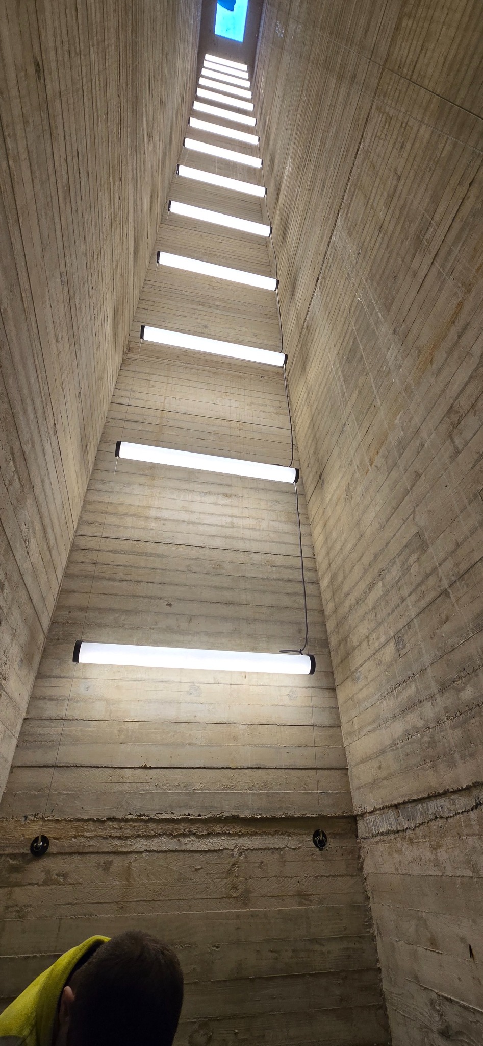 Betonowy szyb z szeregiem lamp liniowych LED, widok z dołu w górę. Minimalistyczne oświetlenie w surowym wnętrzu, z fragmentem osoby w dolnym rogu.