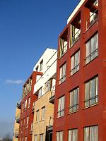 Budynek wielorodzinny z elewacją wentylowaną w kolorze czerwonym i beżowym, widoczne okna z białymi ramami i fragment błękitnego nieba.