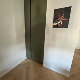 Wbudowana, oliwkowozielona szafa z dwiema pionowymi szczelinami zamiast uchwyt&oacute;w, na ścianie obraz z psem w kr&oacute;lewskim kostiumie, parkiet ułożony w jodełkę.