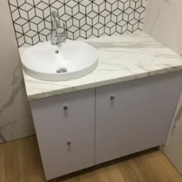 Nowoczesna łazienka z białą, okrągłą umywalką nablatową, chromowaną baterią, szafką w kolorze gołębim, blatem imitującym marmur oraz geometryczną mozaiką na ścianie.