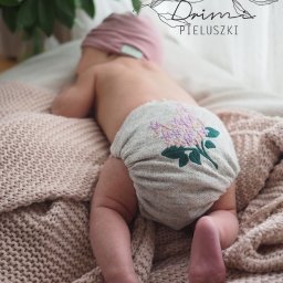 KAAN Aleksander Wadas - Niemowlę leżące na brzuchu na beżowym kocu, ubrane w szarą pieluszkę wielorazową z haftowanym motywem kwiatowym w odcieniach fioletu i zieleni.