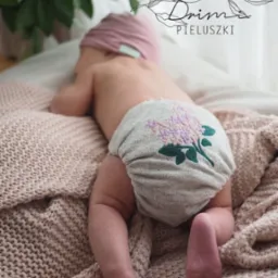 Niemowlę leżące na brzuchu na beżowym kocu, ubrane w szarą pieluszkę wielorazową z haftowanym motywem kwiatowym w odcieniach fioletu i zieleni.