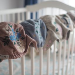 KAAN Aleksander Wadas - Rząd wielorazowych pieluszek wełnianych z haftowanymi kwiatowymi wzorami, zawieszonych na białym łóżeczku dziecięcym, z widoczną metką producenta na pierwszej pieluszce.