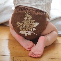 KAAN Aleksander Wadas - Dziecko w brązowej, wełnianej pieluszce wielorazowej z haftowanym, kremowym motywem kwiatowym, siedzące na podłodze. Widoczne małe stopy i fragment białej bluzeczki. W tle jasne, rozmyte tło.