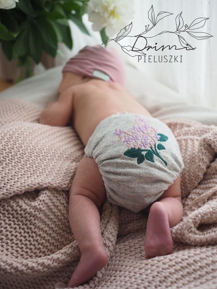 Niemowlę leżące na brzuchu na beżowym kocu, ubrane w szarą pieluszkę wielorazową z haftowanym motywem kwiatowym w odcieniach fioletu i zieleni.