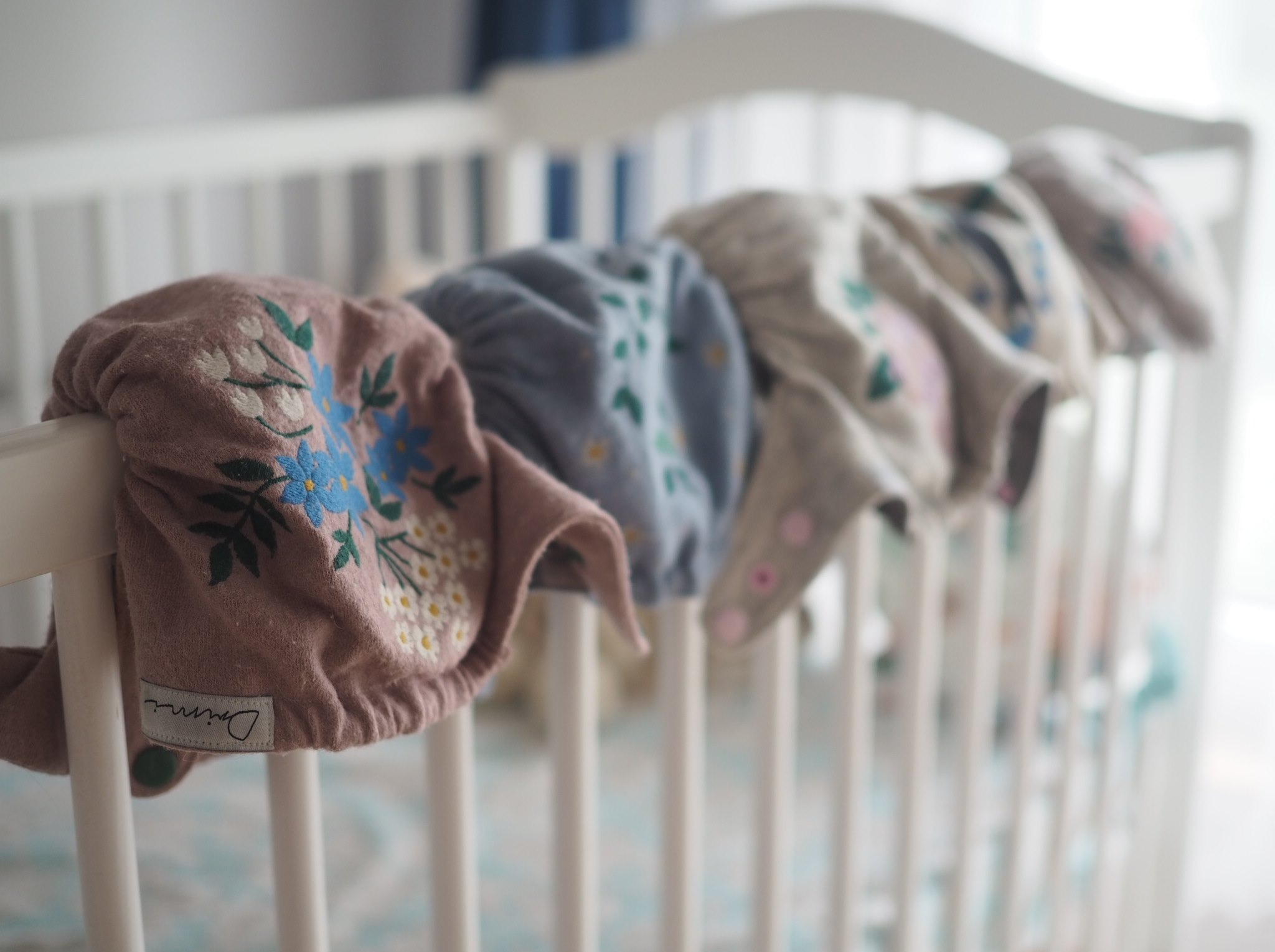 Rząd wielorazowych pieluszek wełnianych z haftowanymi kwiatowymi wzorami, zawieszonych na białym łóżeczku dziecięcym, z widoczną metką producenta na pierwszej pieluszce.