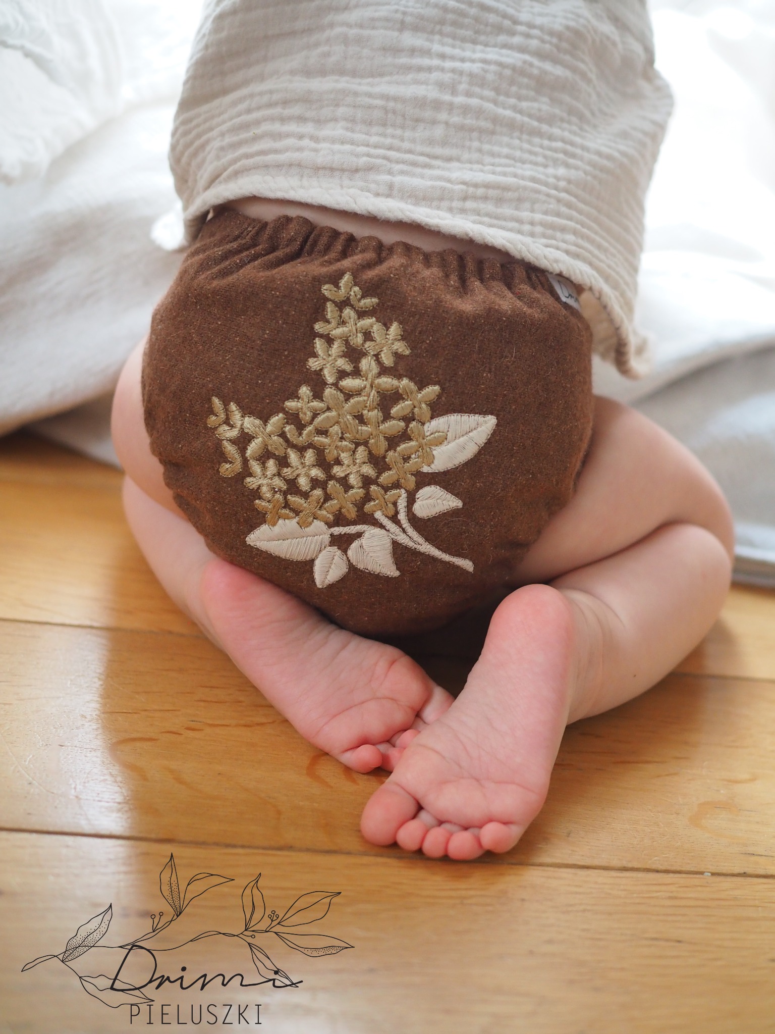 Dziecko w brązowej, wełnianej pieluszce wielorazowej z haftowanym, kremowym motywem kwiatowym, siedzące na podłodze. Widoczne małe stopy i fragment białej bluzeczki. W tle jasne, rozmyte tło.