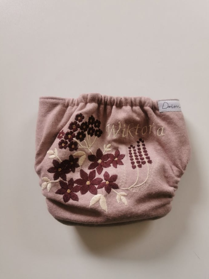 Różowa wełniana pieluszka wielorazowa z haftowanym imieniem 'Wiktora' i motywem kwiatowym, leżąca na gładkim, jasnym tle.