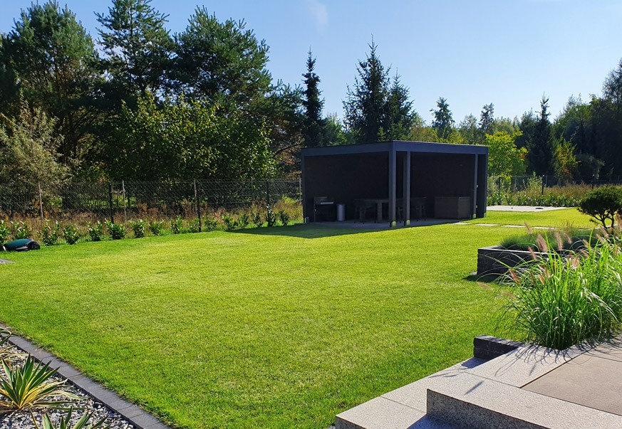 Zadbany trawnik z robotem koszącym, nowoczesna altana z grillem i meblami ogrodowymi, ozdobne nasadzenia traw i krzewów. Całość w słoneczny dzień.