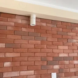 Ściana wykończona cegłą klinkierową w odcieniach czerwieni i brązu, oświetlona dwoma cylindrycznymi lampami sufitowymi w kolorze białym, z widocznym włącznikiem światła na dole ściany.