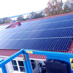 KAJ-TECH SPÓŁKA Z OGRANICZONĄ ODPOWIEDZIALNOŚCIĄ - Instalacja paneli słonecznych na dachu z czerwonej dachówki, widok z podnośnika koszowego, w tle krajobraz z jeziorem.