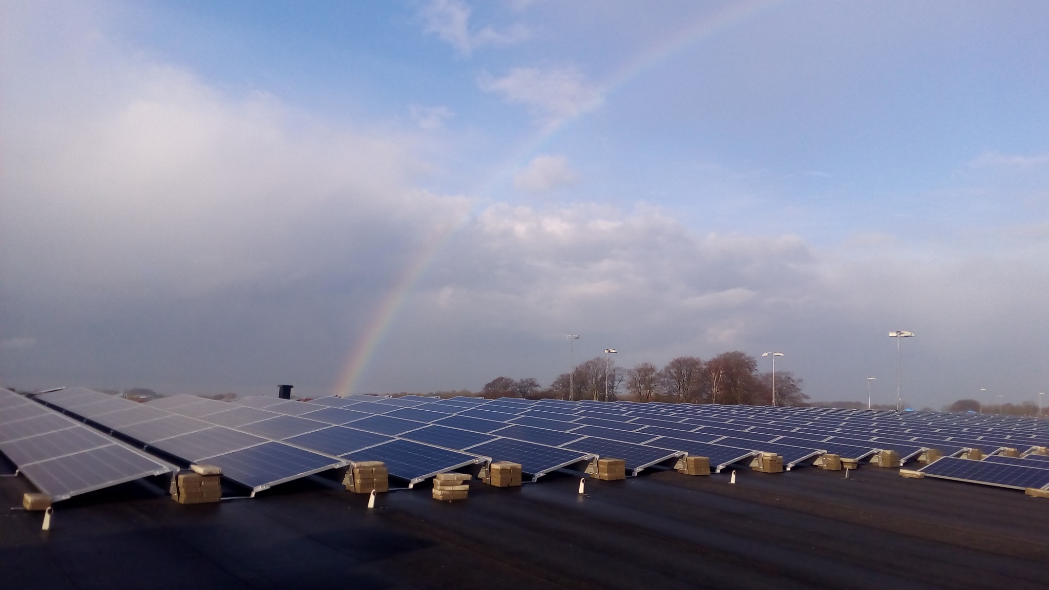 Rozległa instalacja paneli słonecznych na dachu, zabezpieczona cegłami, z widoczną tęczą na pochmurnym niebie w tle.