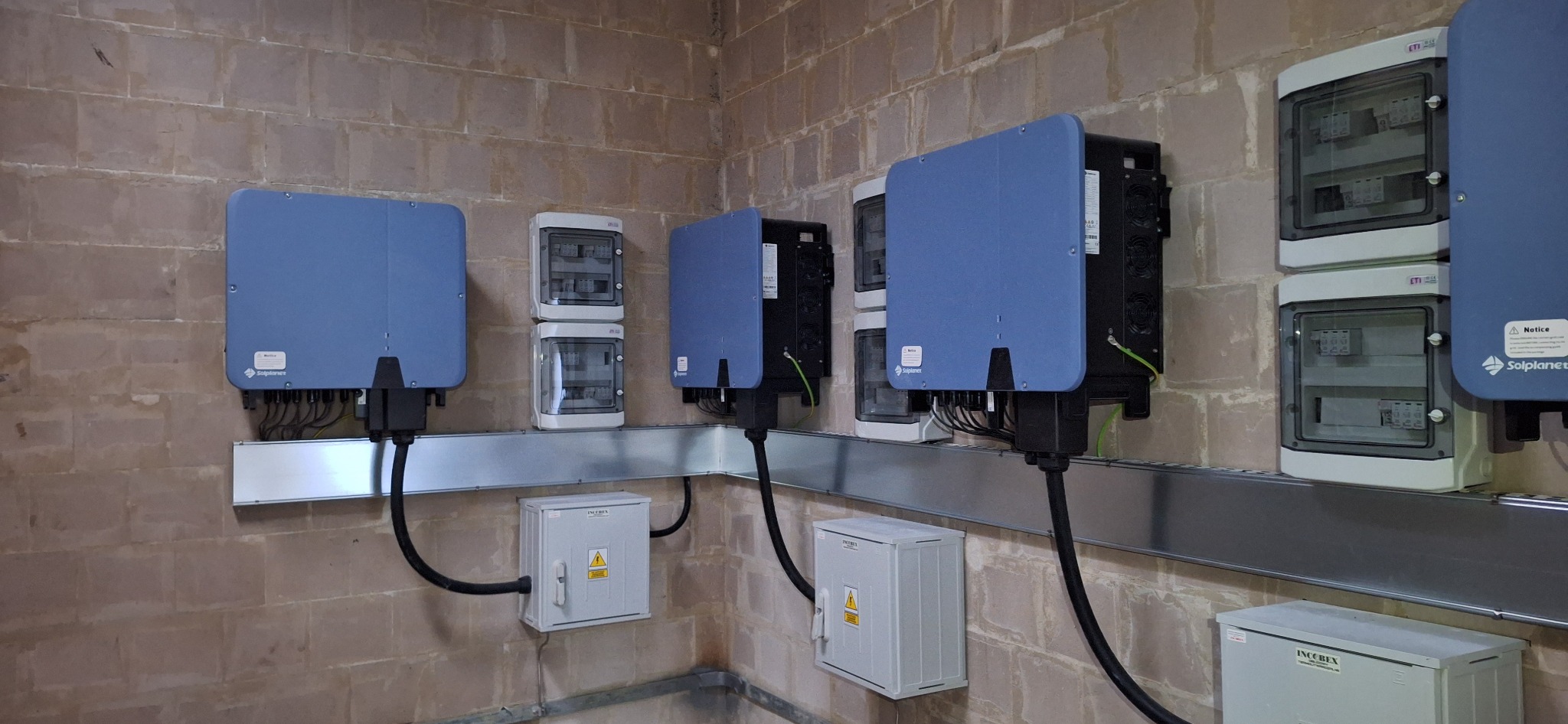 Instalacja fotowoltaiczna w pomieszczeniu technicznym: falowniki Solplanet i skrzynki rozdzielcze ETI zamontowane na szynie, dodatkowe zabezpieczenia w obudowach INCOBEX na ceglanej ścianie.