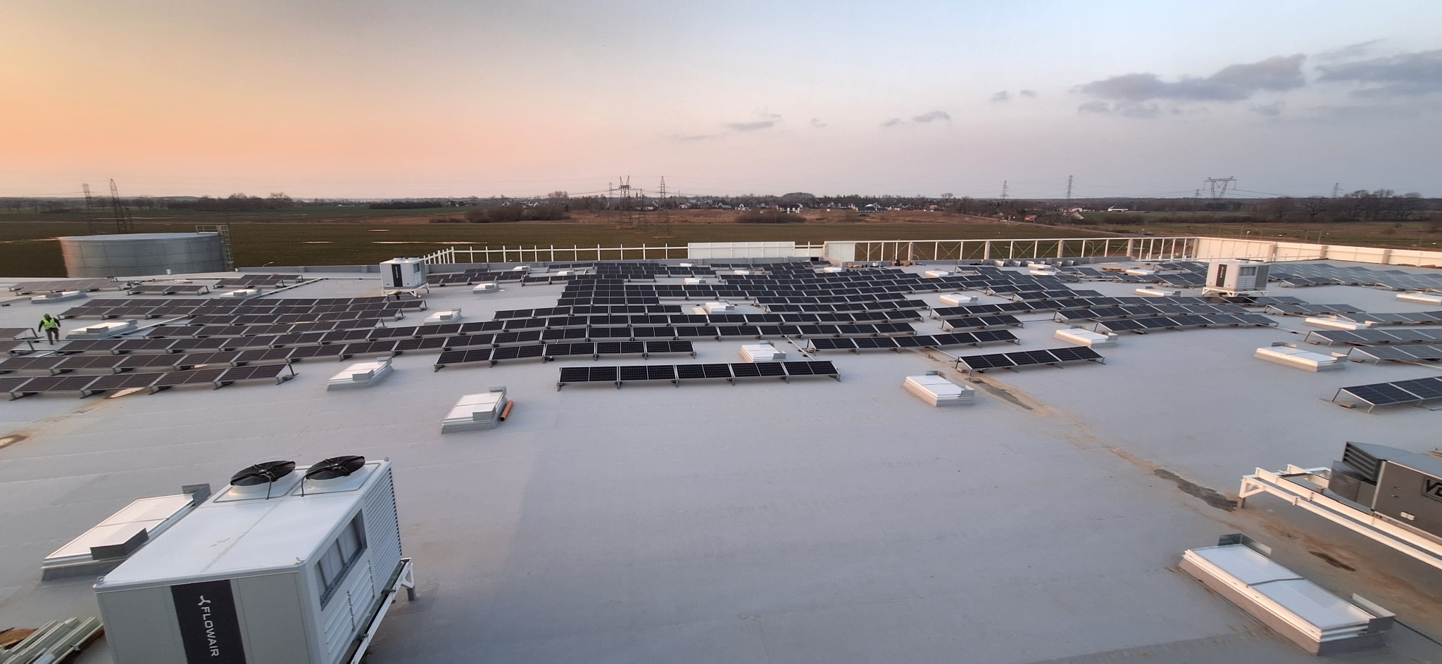 Rozległa instalacja paneli słonecznych na płaskim dachu budynku przemysłowego, widoczny pracownik w odblaskowej kamizelce, zbiornik na wodę i wiejskie tereny w tle pod zachodzącym słońcem.