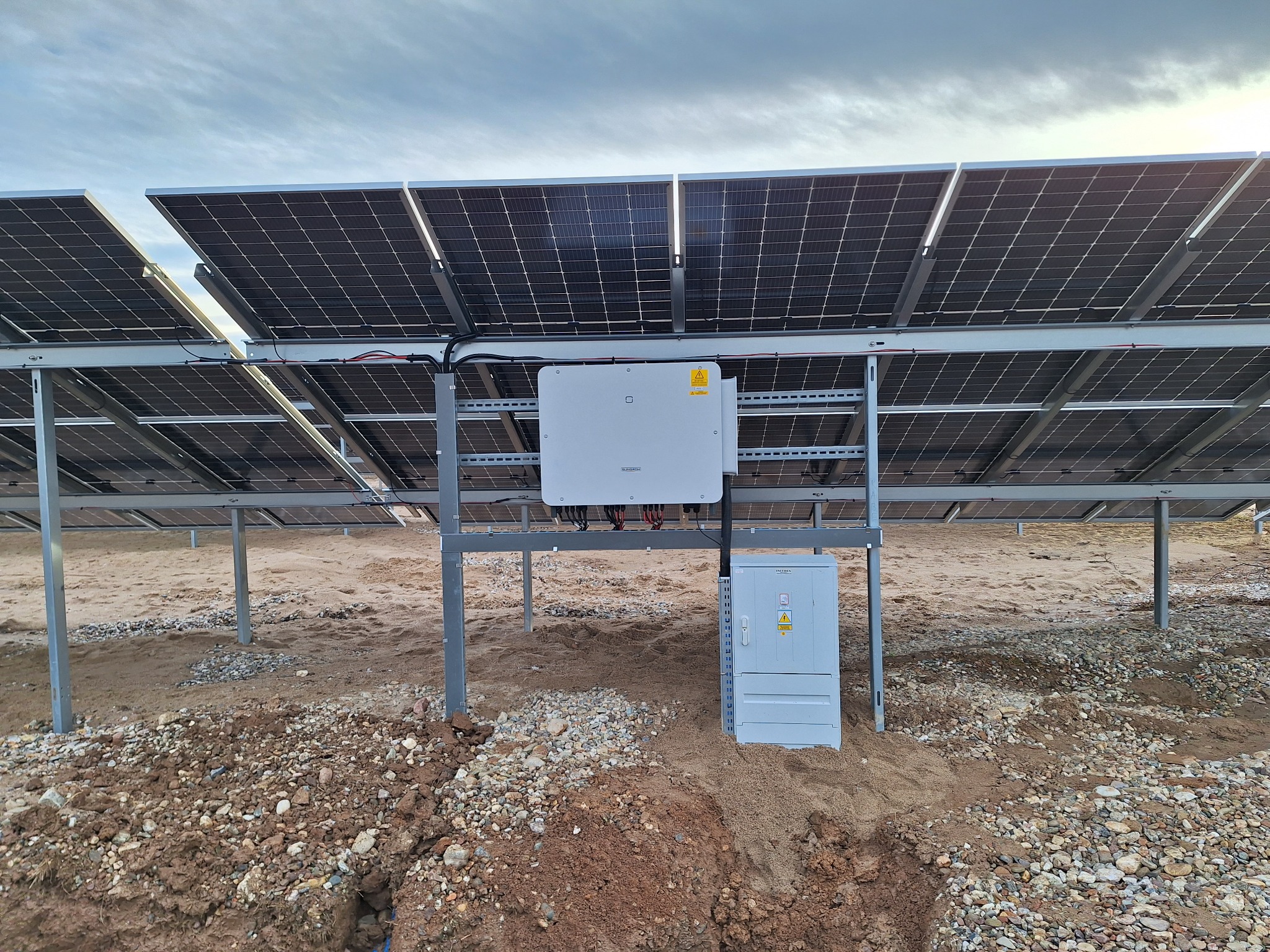 Widok od spodu na panele fotowoltaiczne zamontowane na metalowej konstrukcji, z białym inwerterem Huawei i szarą skrzynką elektryczną na piaszczystym podłożu z kamieniami.