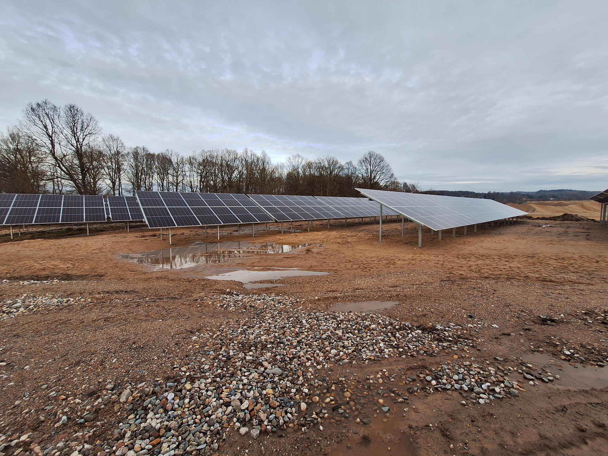 Rzędy paneli słonecznych na ziemi z kamieniami i kałużami, pochmurne niebo w tle.