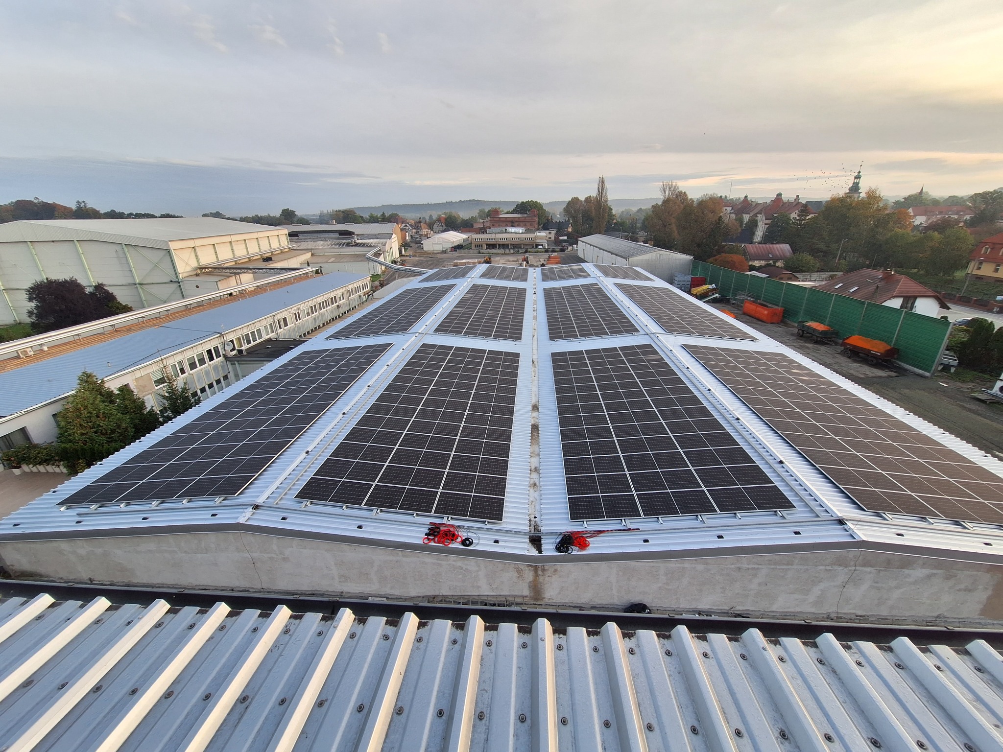 Rozległa instalacja solarna na dachu budynku przemysłowego, widok z góry, z widocznymi przewodami i elementami montażowymi, w tle zabudowania miejskie pod pochmurnym niebem.