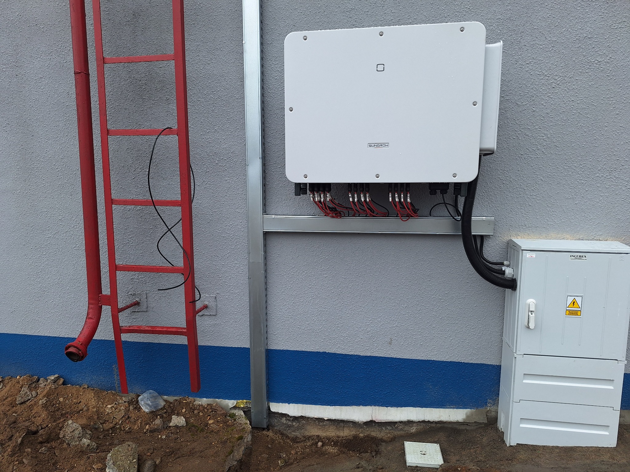 Biała skrzynka inwertera firmy Sungrow zamontowana na szarej ścianie budynku z widocznymi przewodami, obok skrzynka zabezpieczeń INCOBEX, z czerwoną rurą spustową i drabinką po lewej stronie.