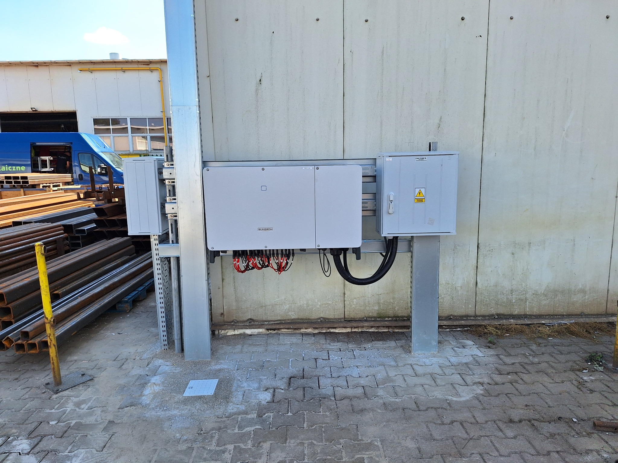 Zamontowany inwerter fotowoltaiczny Sungrow z okablowaniem i skrzynką zabezpieczeń na zewnątrz budynku przemysłowego, widoczne elementy konstrukcyjne i transportowy bus w tle.