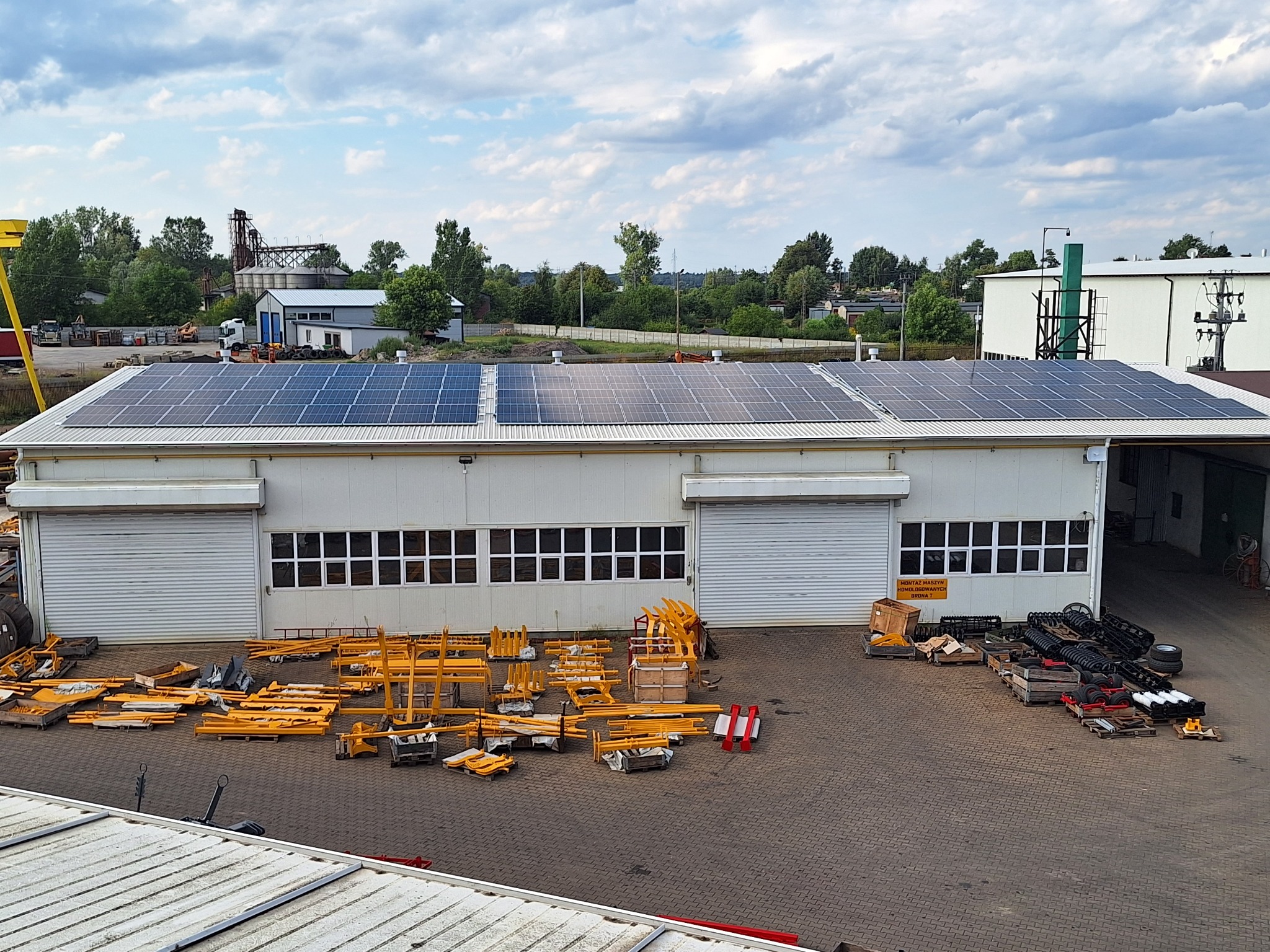 Budynek przemysłowy z panelami słonecznymi na dachu, przed budynkiem rozłożone żółte elementy konstrukcyjne i czarne części maszyn.