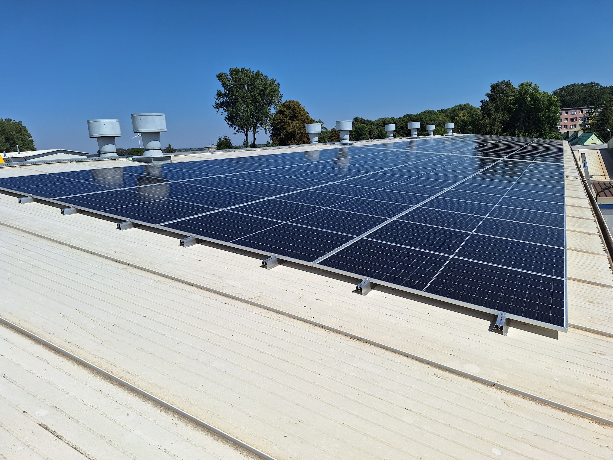 Instalacja fotowoltaiczna na dachu budynku przemysłowego z widocznymi elementami wentylacji i turbiną wiatrową w tle, jasne niebo.