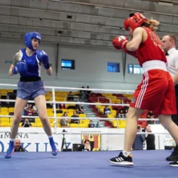 Dwie zawodniczki boksujące na ringu, jedna w niebieskim stroju i kasku, druga w czerwonym, z sędzią w tle i publicznością na trybunach.