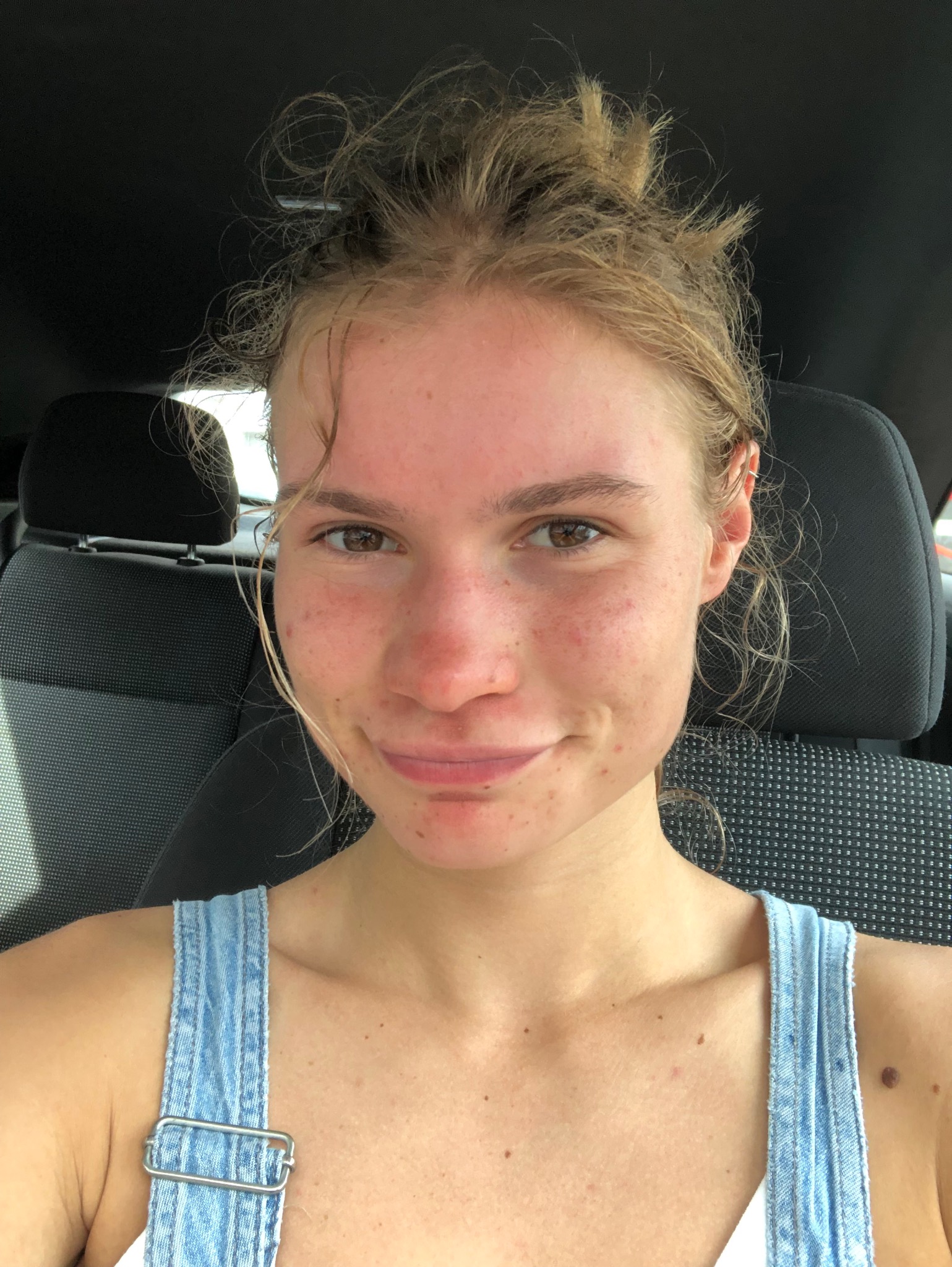 Portret uśmiechniętej, młodej kobiety z piegami, siedzącej w samochodzie, z włosami upiętymi w niedbały kok i ubrana w dżinsowy kombinezon.