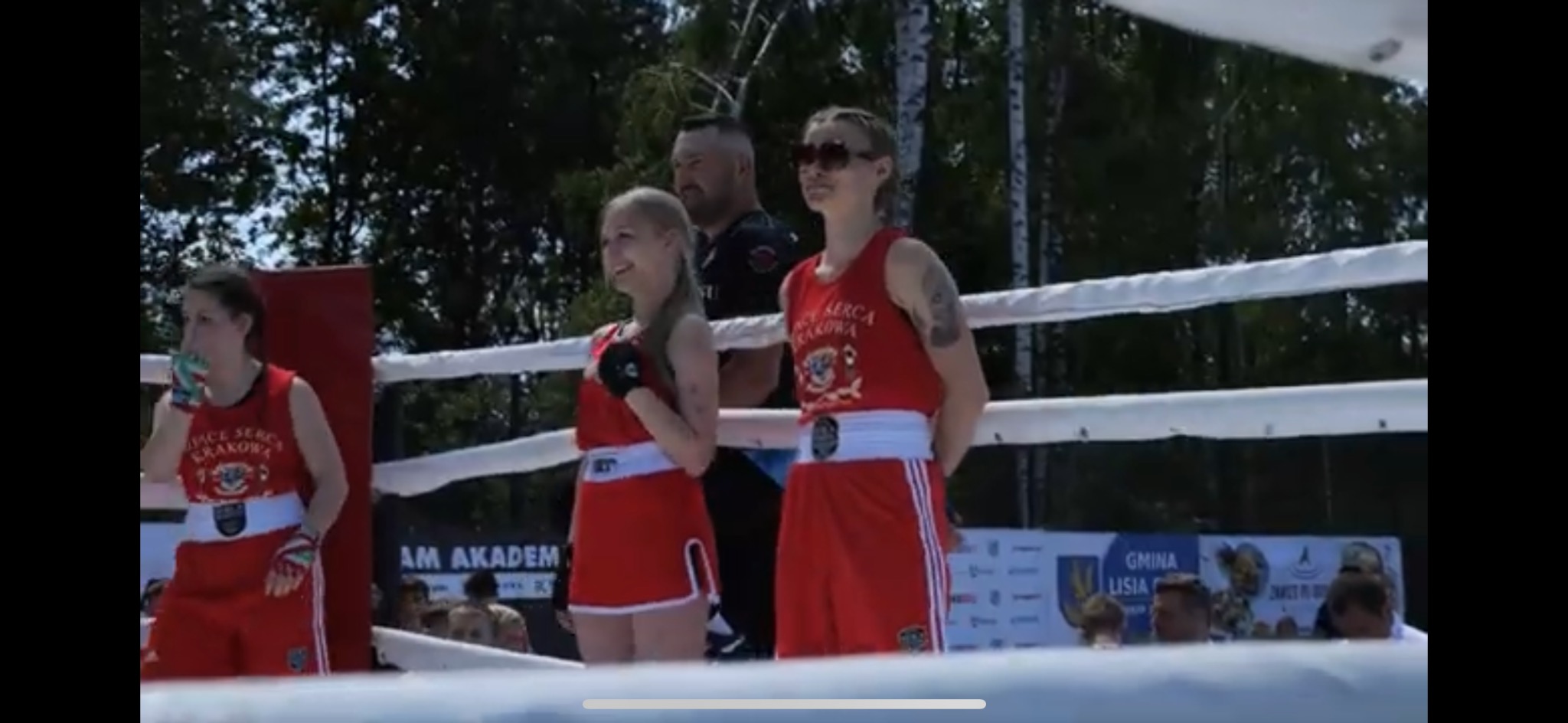 Trzy zawodniczki boksu w czerwonych strojach z logo drużyny 'Serce Serca Krakowa' stoją na ringu przed publicznością, z trenerem w tle.