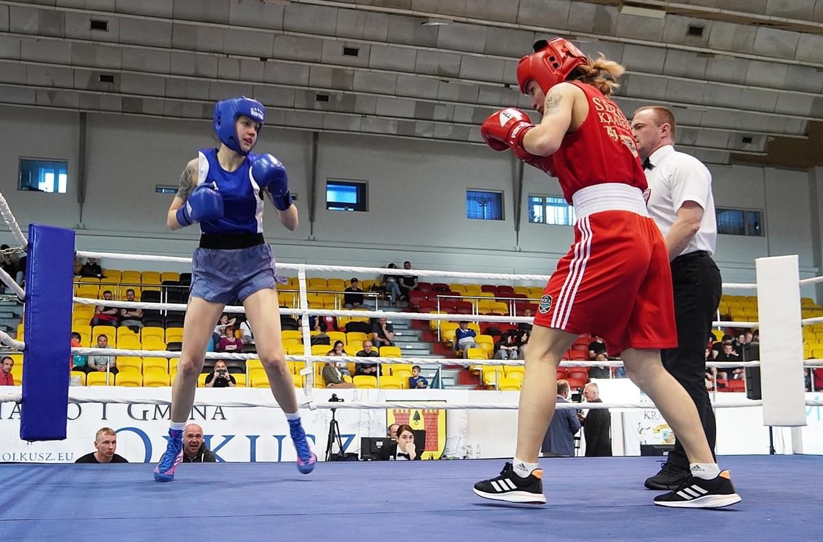 Dwie zawodniczki boksujące na ringu, jedna w niebieskim stroju i kasku, druga w czerwonym, z sędzią w tle i publicznością na trybunach.