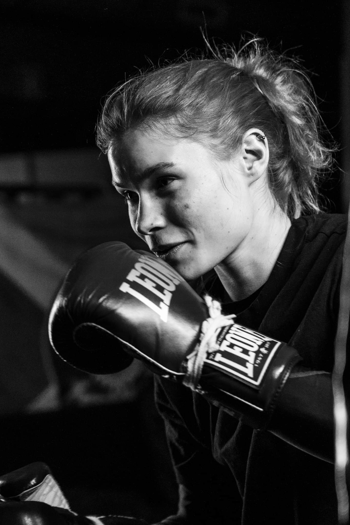 Czarno-białe zdjęcie kobiety z kolczykiem w uchu, w bokserskich rękawicach marki Leone, spoglądającej w lewo z lekkim uśmiechem.