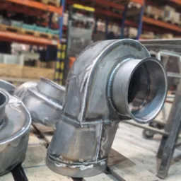 Metalowe kolanka spawane z widocznymi śladami spawania, ułożone na drewnianej palecie pomalowanej na różowo, w tle magazyn z regałami.