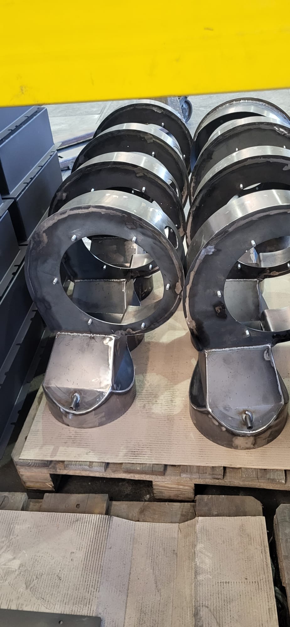 Dwa rzędy spawanych, metalowych elementów konstrukcyjnych z okrągłymi otworami i podstawami, leżące na drewnianej palecie w warsztacie.