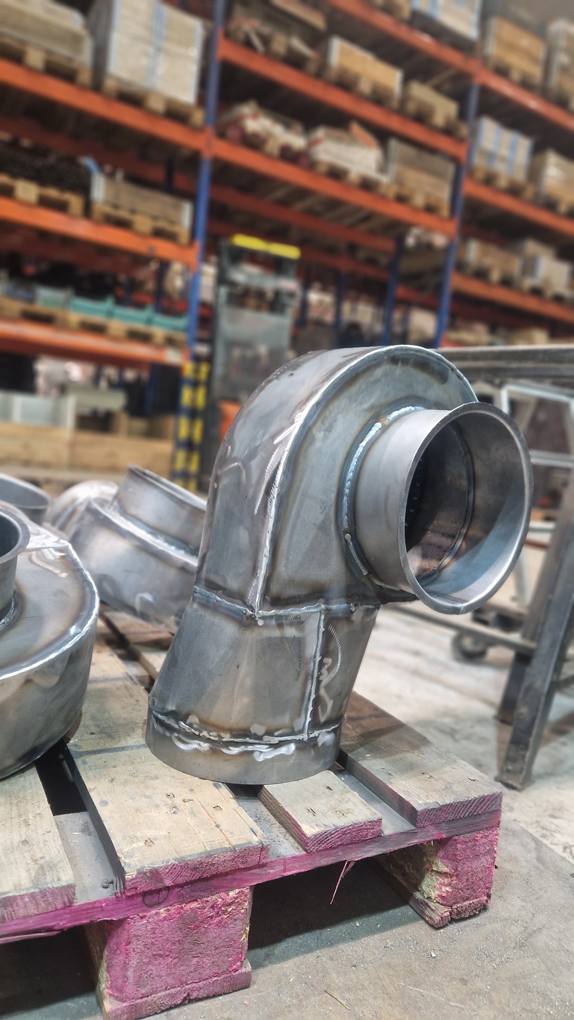 Metalowe kolanka spawane z widocznymi śladami spawania, ułożone na drewnianej palecie pomalowanej na różowo, w tle magazyn z regałami.